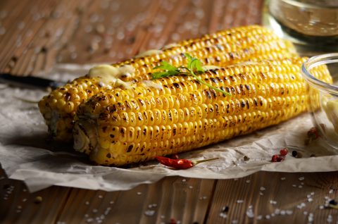 Kitchen table with grilled sweet corn cob under melting butter and greens on baking paper